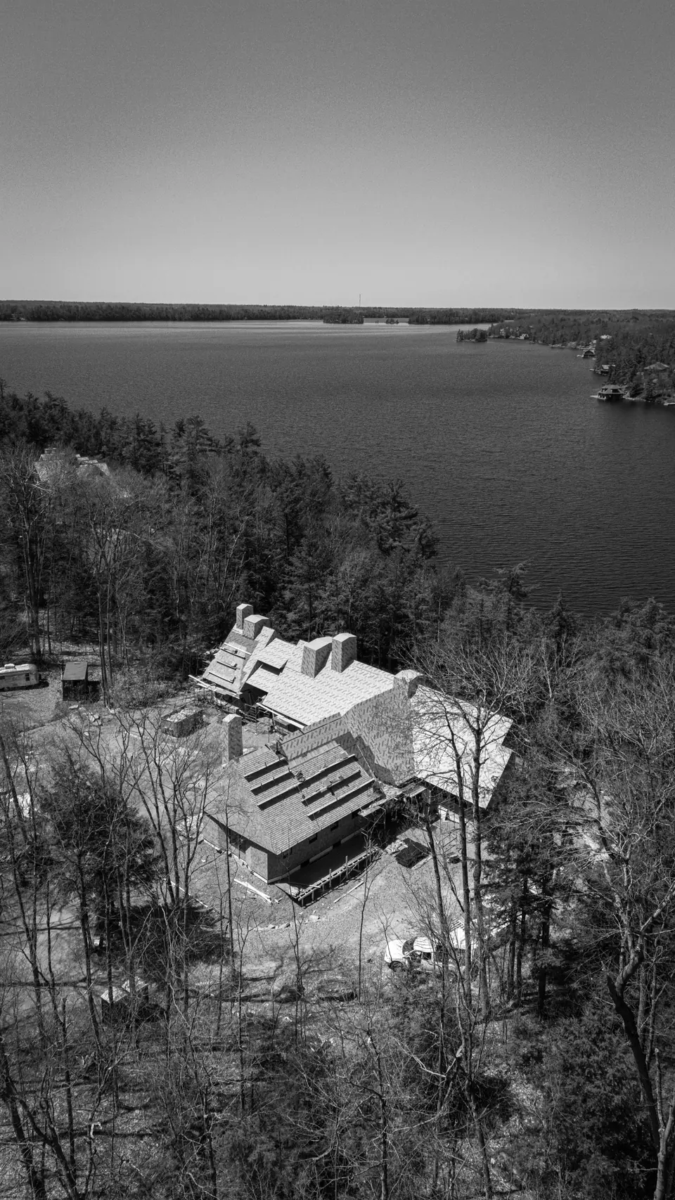 Muskoka Modern Shingle Style house under construction on Lake Muskoka, Ontario: aerial view of the cedar-shingle residence among the pines, with the wide lake and open sky beyond