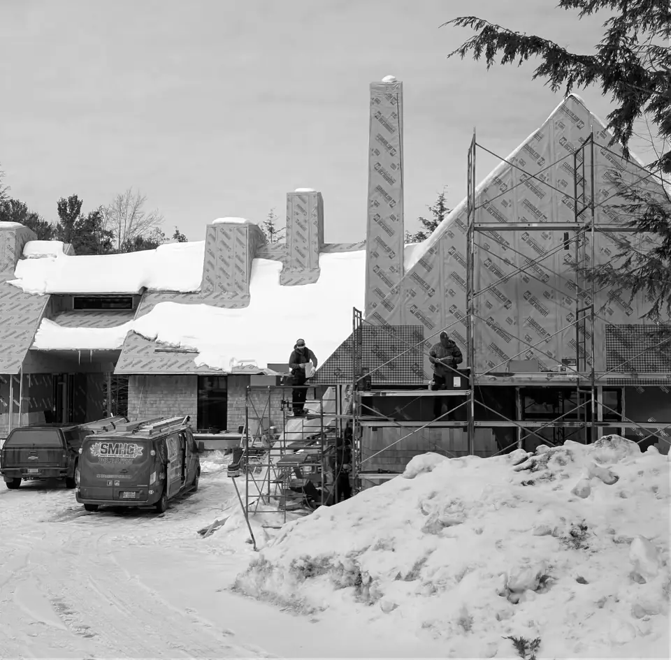 Muskoka Modern Shingle Style house under construction on Lake Muskoka, Ontario: winter on site: snow on the roofs, the house in house wrap with tall chimneys, scaffolding, builders and work vans